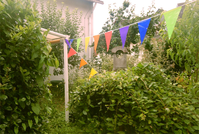 Ein Garten mit grünen Büschen. Bunte Wimpel sind quer gespannt. 